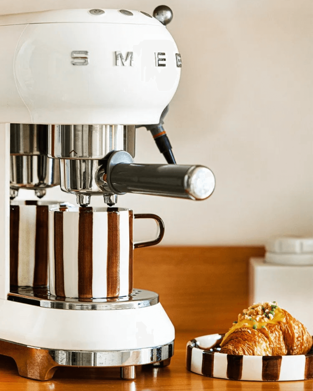 Smeg espresso machine with a striped ceramic cup and croissant on a wooden countertop.