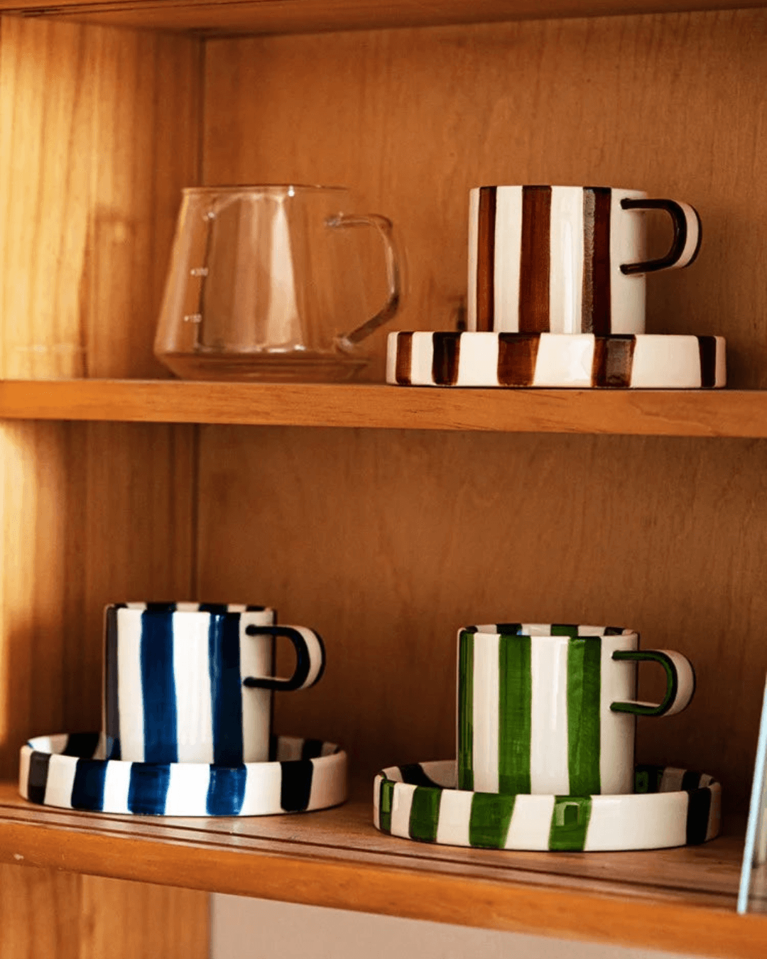Ceramic coffee mug set with retro striped design, featuring green, blue, and brown mugs with matching saucers on a wooden shelf.