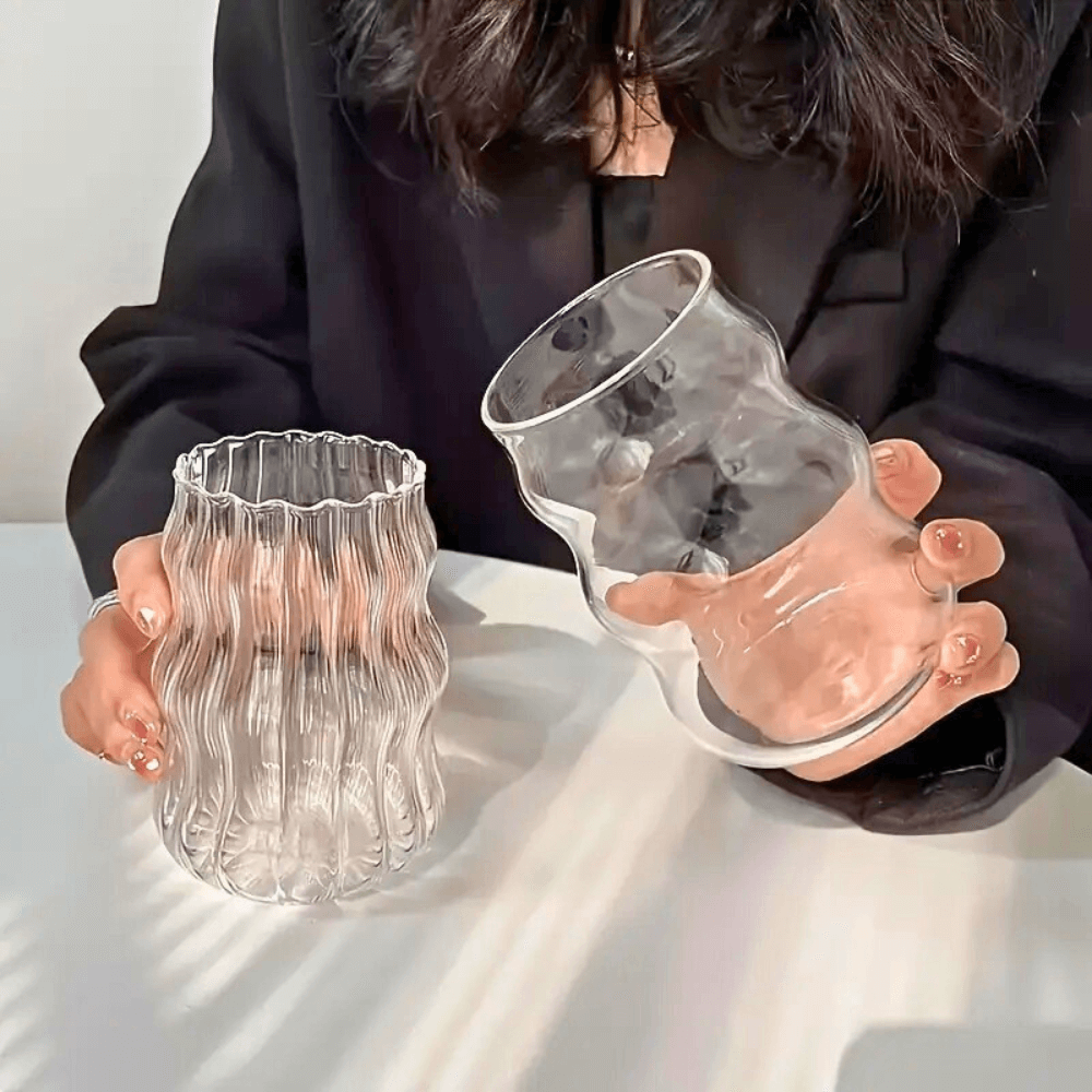 Two heat-resistant glass tumblers in hand, featuring modern wavy and smooth designs for iced drinks.