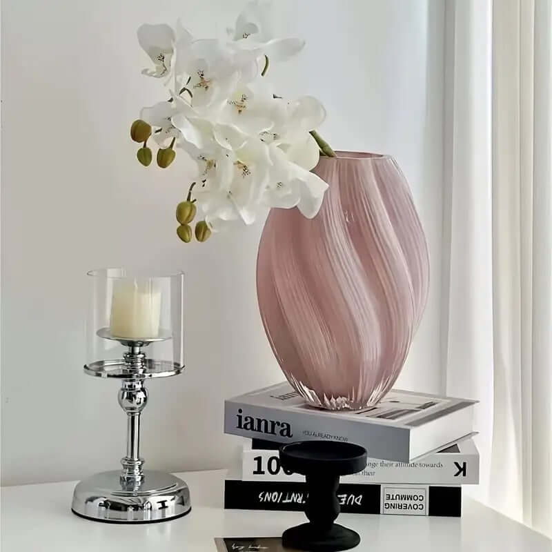 Elegant spiral glass flower vase with white orchids on books beside a candle holder.