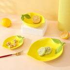 Lemon-shaped ceramic plate, bowl, and dish set in yellow featuring fruit-inspired design on a bright table.