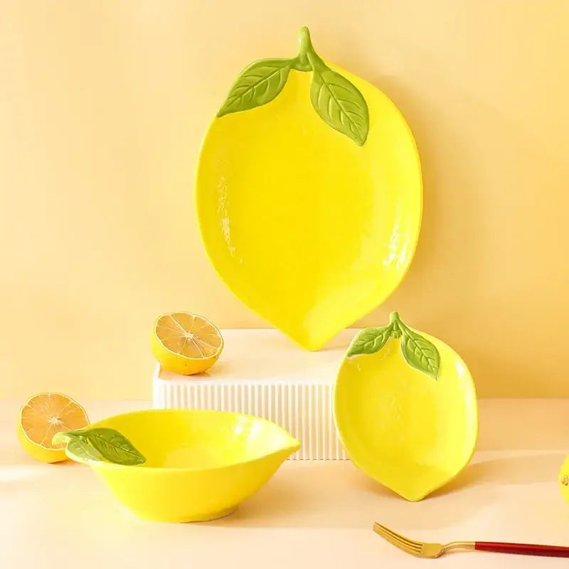Lemon-shaped ceramic tableware set including plate, bowl, and dish in bright yellow with green leaf accents.