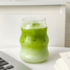 500ml wavy matcha glass with iced matcha drink on a white table beside a laptop and a book.