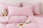 Cozy pink gingham bedding set with plush pillows, teddy bear, and mug on a soft bed, perfect for a serene bedroom.