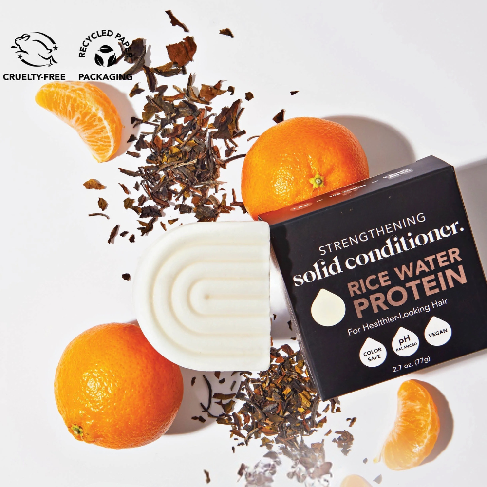 Strengthening solid conditioner with rice water protein, surrounded by oranges and tea leaves, cruelty-free and recyclable packaging.