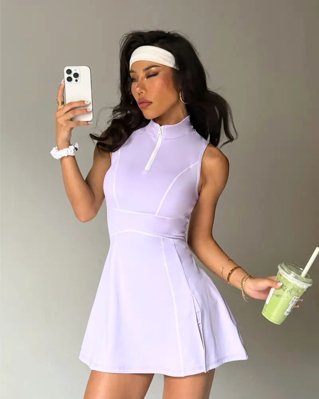 Stylish woman in a lavender padel tennis dress with zip polo neck, holding a drink and taking a selfie.