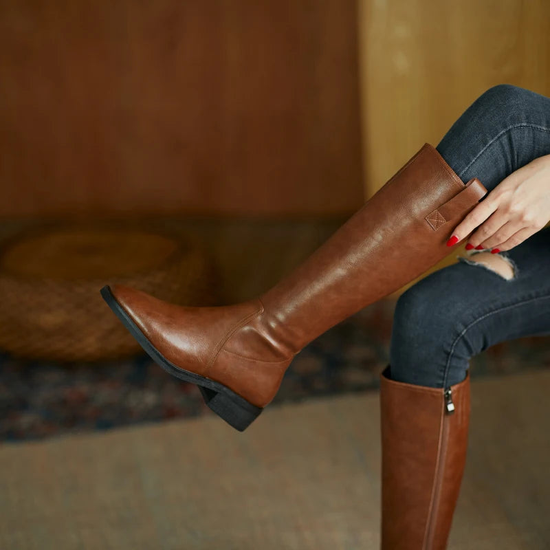 Elegant women’s knee-high leather boots in brown, featuring a pointed toe and chunky heel, styled with denim pants.