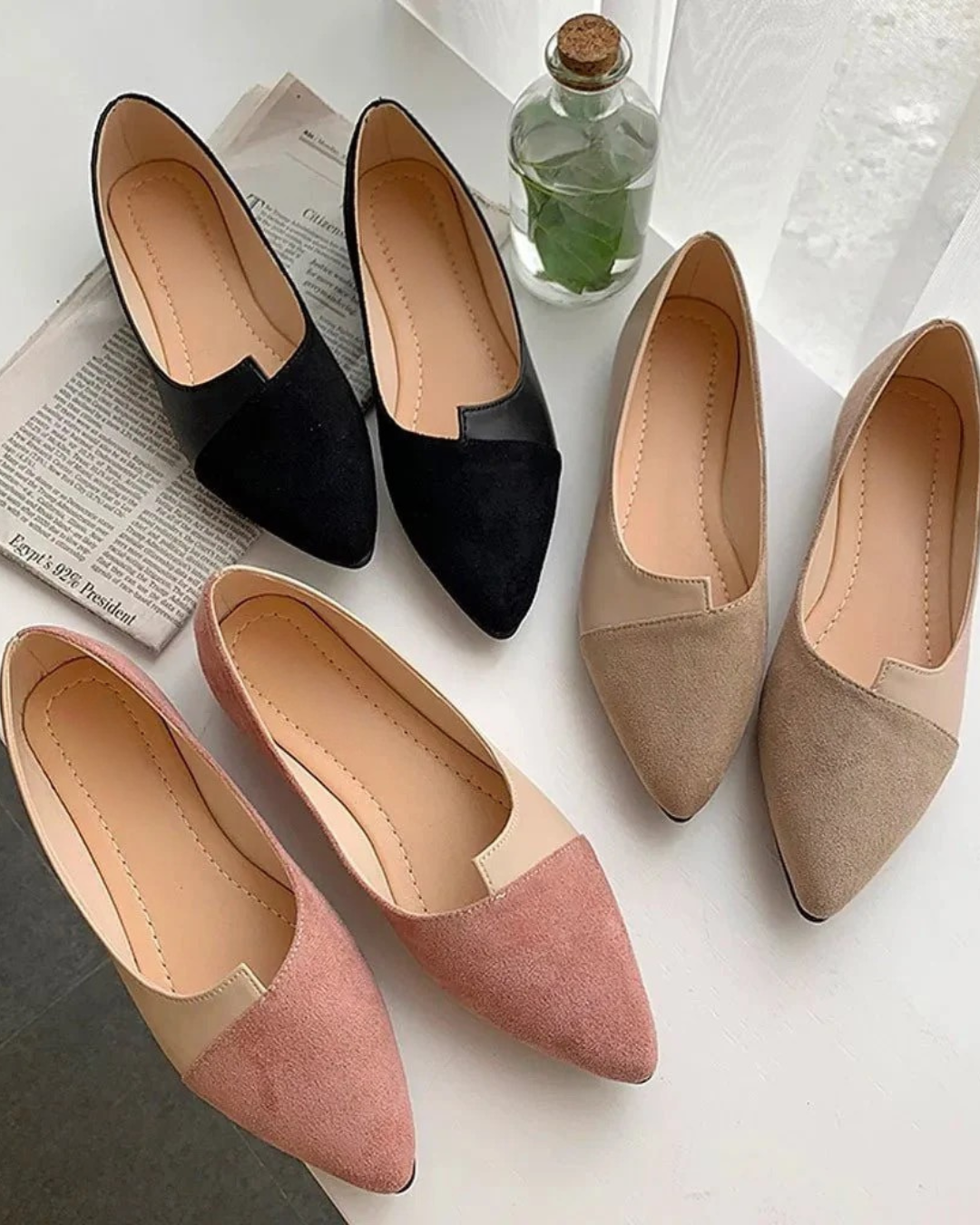 Pointed toe flats in black, beige, and pink color options displayed on a table with greenery and a newspaper.